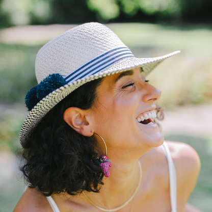 A woman outdoors wearing a white woven sun hat with blue stripes and a dark blue pom-pom accent, a white camisole top, and gold hoop earrings.