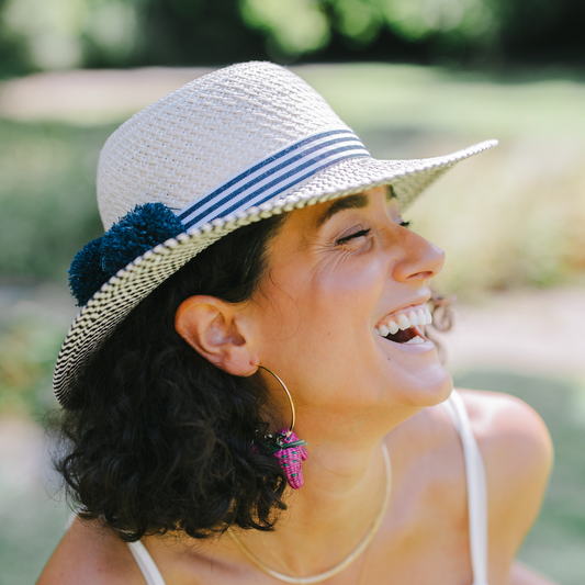 A woman outdoors wearing a white woven sun hat with blue stripes and a dark blue pom-pom accent, a white camisole top, and gold hoop earrings.