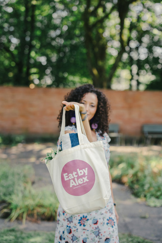 Woman holding a tote bag with 'Eat by Alex' logo in an outdoor setting