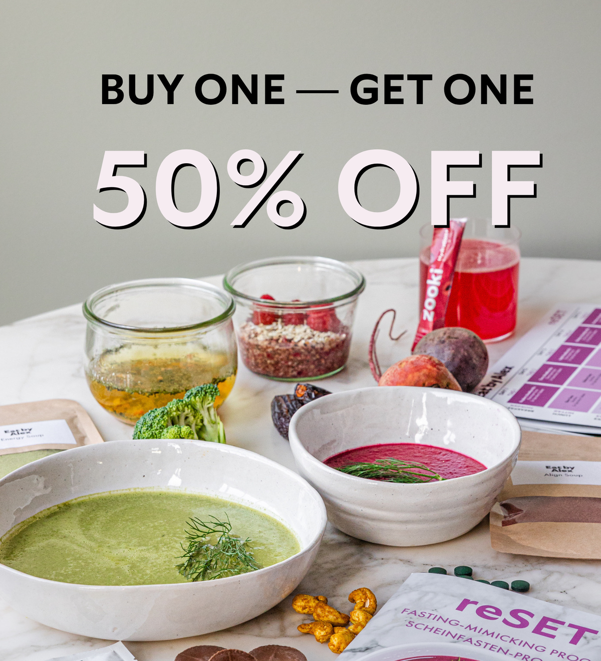 A promotional sale graphic with the text 'BUY ONE - GET ONE 50% OFF' over a styled food-prep scene. White bowls containing green and pink soups are arranged on a marble surface with jars, produce, and a pink bottle in the background.
